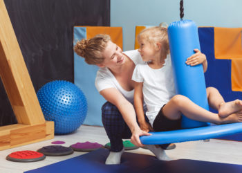 Young physiotherapist seesawing the girl on a blue swing